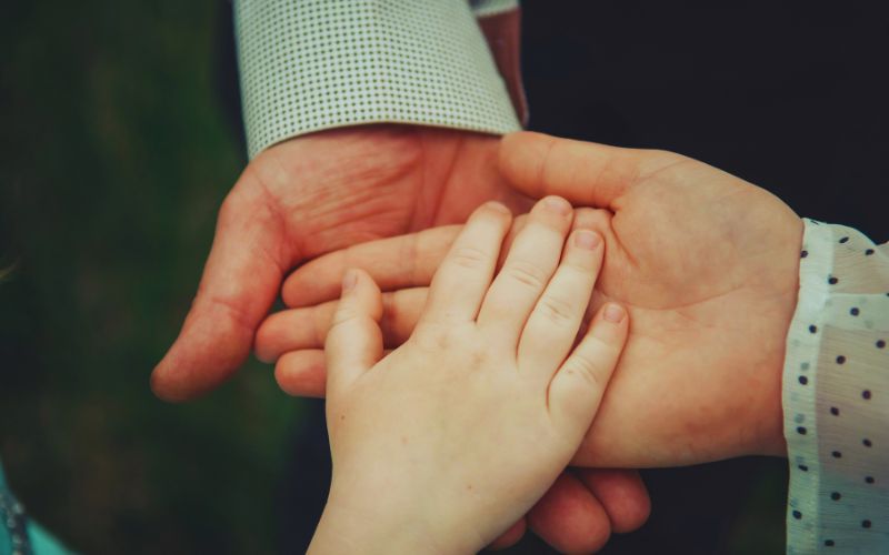 Close-up of two adult hands gently holding a child's hand, symbolising support in parenting after separation. The adults wear long-sleeved, patterned shirts, with the child's hand resting on top against a softly blurred green background.