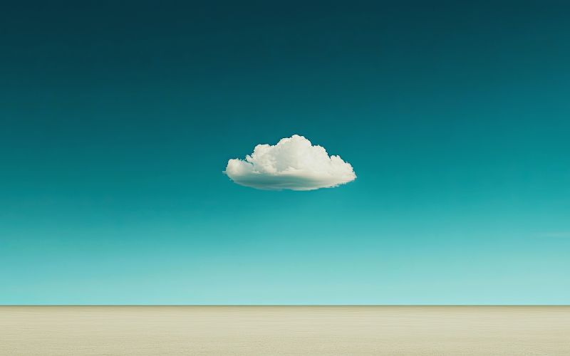 A single fluffy white cloud floats in a clear blue sky above a flat, empty beige landscape.