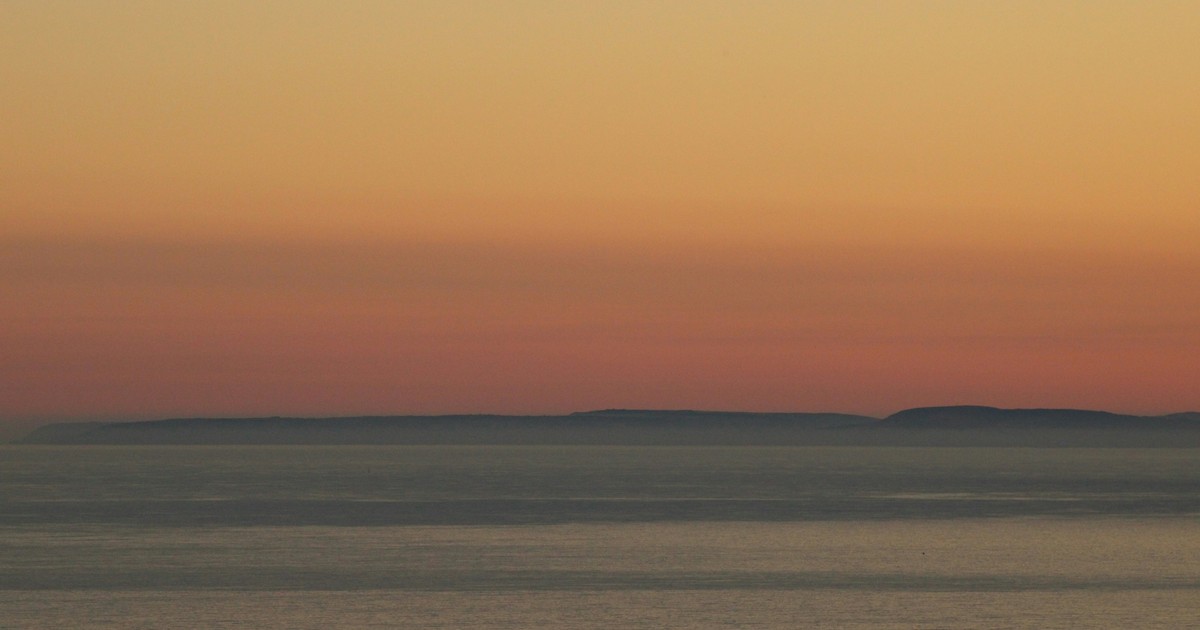 A tranquil ocean scene at sunset, with calm water in the foreground and a distant landmass under a gradient sky of orange, pink, and yellow hues.